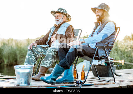 Großvater mit erwachsenen Sohn angeln zusammen auf der hölzernen Pier im Morgenlicht. Blick von der Seite des Sees Stockfoto