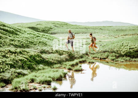 Mann und Frau Wandern in der Nähe des Sees in die Berge, die Landschaft Blick auf die grüne Wiese mit See während der sonnigen Tag Stockfoto