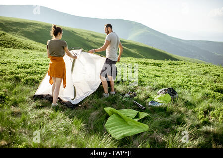 Junges Paar einrichten das Zelt auf der grünen Wiese, Reisen hoch in die Berge bei Sonnenuntergang Stockfoto
