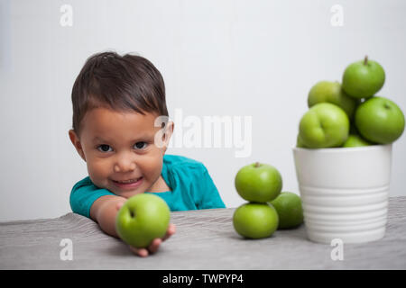 Kleiner Junge mit einem frischen grünen Apfel in der Hand und bietet es oder es von seinem Haufen von Obst. Stockfoto