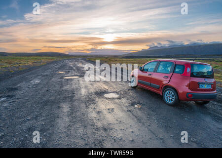 Sonnenuntergang auf unbefestigte Straße mit Schlaglöchern in Island, roten Fiat Panda entlang der Straße, während die Sonne hinter den Bergen ist. Stockfoto