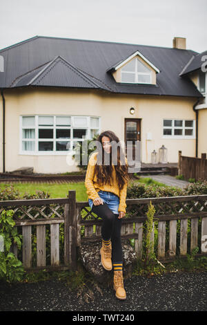 Junge glücklich brünette Frau in orange Pullover und Stiefel sitzen auf Zaun mit Hintergrund der traditionellen skandinavischen Hausfassade. Herbst. Haus A Stockfoto