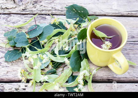 Linden Tee in eine gelbe Schale steht in der Nähe von Linden Blumen vor dem Hintergrund des alten Boards. Kräutertee mit Linden Blumen. Stockfoto