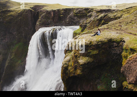 Frau sitzt auf der Kante mit atemberaubenden Blick auf fagrifoss Wasserfall in Island. Lakagigar Wahrzeichen in Skaftafell National Park. Stockfoto