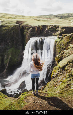 Curly Frau auf fagrifoss Wasserfall in Island suchen. Ansicht von der Rückseite, Wind im Haar, gute sonnige Sommerwetter. Stockfoto