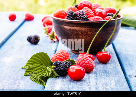 Saftige frische leckere Beeren, Kirschen, Himbeeren, Maulbeeren in einer Schüssel auf Blau Boards auf einem Hintergrund von grünem Gras. Close-up Stockfoto