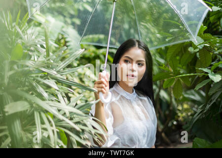 Asiatische Frau mit Schirm aus transparentem Kunststoff in den regnerischen Tag bei Aufenthalt im Freien bei tropischen Wald mit grünen Blättern Stockfoto