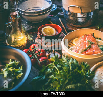 Gesundes Essen und Kochen. Lachsfilet in Bambus Steamer am Küchentisch mit sauber Zutaten: frische Kräuter, Gemüse, Zitronensaft und Öl Flasche. Stockfoto