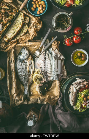 Tisch mit gesicherten dorado Fisch, Salat und Brot auf dunklen rustikalen Tabelle, Ansicht von oben. Mediterranes Mittagessen oder Abendessen. Gesunde Ernährung Stockfoto