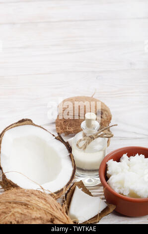 Hintergrund der Kokosnuss, Kokosnussschalen, Öl in Ton Schüssel und Glas auf weissem Holztisch Stockfoto