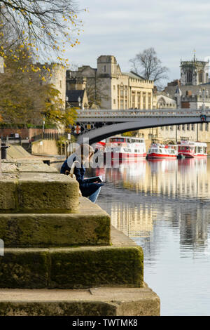 Ruhigen malerischen Lage am Fluss von Lendal Brücke (Boote im Fluss Ouse & Frau Lesung wider am Ufer) - York, North Yorkshire, England, UK. Stockfoto