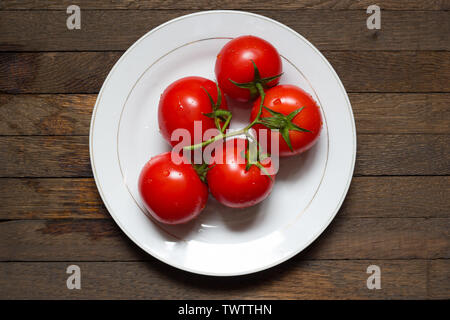 Rote Tomaten am Zweig auf weiße Platte auf nassem Holz- Tabelle. Die mittlere Stellung. Ansicht von oben. Nahaufnahme. Wassertropfen auf frischem Gemüse. Gesunde Ernährung m Stockfoto