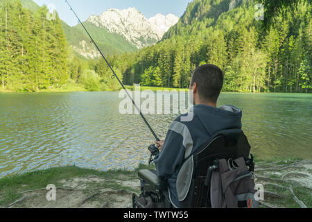 Junge Mann in einem Rollstuhl Angeln am See an einem sonnigen Tag, mit den Bergen im Rücken Stockfoto