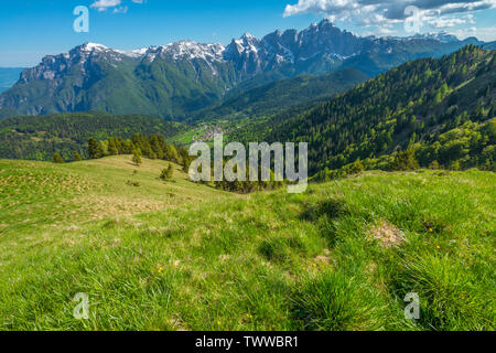 Blick auf die Berge Stadt unten im Tal von einem grünen Almwiese. Üppig bewachsenen Berghang in den italienischen Alpen im Juni. Stockfoto