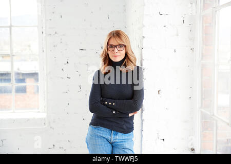 Portrait von attraktiven Frau mittleren Alters tragen Rollkragen Pullover und Jeans beim Entspannen am Fenster und Blick auf Kamera. Stockfoto