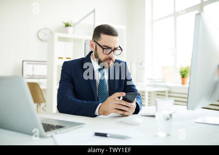 Ernsthafte konzentrierte junge Bartgeier Geschäftsmann in Anzug am Schreibtisch im Büro sitzen und lesen SMS auf dem Smartphone Stockfoto