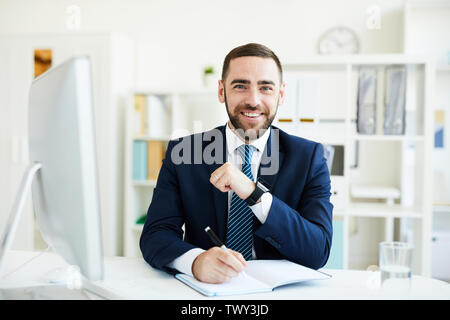 Portrait von Happy Begeistert junge Bartgeier CEO im Anzug am Tisch mit Computer sitzen und schreiben Sie Ziele im Tagebuch Stockfoto
