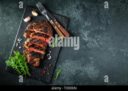 Ribeye Steak in Scheiben, gegrillt mit Pfeffer, Knoblauch, Salz und Thymian auf einer Schiefertafel Schneidbrett auf einem dunklen Stein serviert. Ansicht von oben mit der Kopie Raum Stockfoto