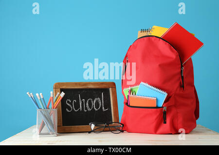 Rucksack mit Lieferungen und Schiefertafel mit Text Schule am Tisch gegen Farbe Hintergrund Stockfoto