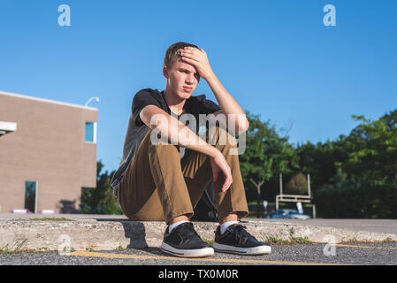 Verärgert Teenager seinen Kopf auf seine Hand, als er auf dem Bürgersteig vor seiner Schule sitzt. Stockfoto