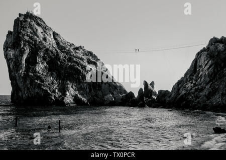 Zwei Menschen laufen an einem Seil Bridge angeschlossen sind Seile zur Sicherheit. Die Brücke ist zwischen zwei Felsen über das Meer ausgestreckt. Russland, der Krim. bw, Toning. Stockfoto