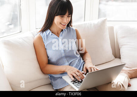 Portrait von wunderschönen Frau eingeben und mit Silber Laptop auf der Couch in helle Wohnung sitzen Stockfoto