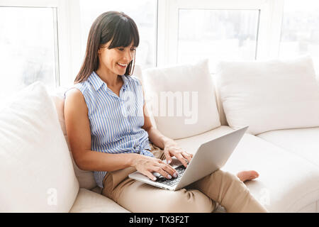 Portrait von fröhlichen Frau 40 s in eleganter Kleidung arbeiten an Silber laptop beim Sitzen auf einem Sofa in helles Apartment Stockfoto