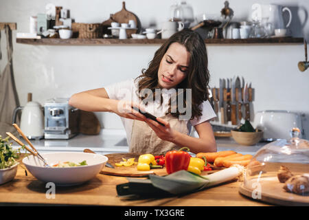 Schöne junge Frau mit Schürze Kochen gesund Salat an der Küche zuhause, mit einem Foto von Lebensmitteln mit Handy Stockfoto