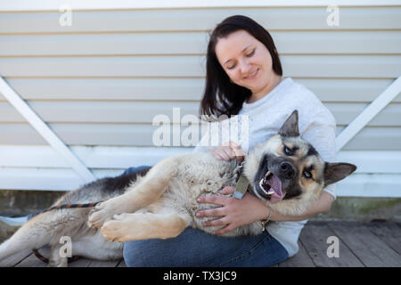 Bild der lächelnde Frau in Jeans und weiße Jacke neben Happy shepherd Welpen gegen weiße Gebäude aus Holz sitzen Stockfoto