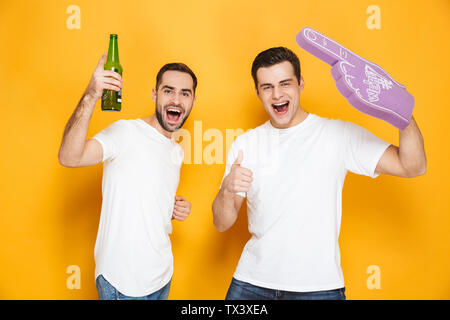 Zwei fröhliche aufgeregt Männer Freunde tragen Leer t-shirts über Gelb Hintergrund isoliert stehen, jubeln mit Schaum Handschuh und Bier Stockfoto