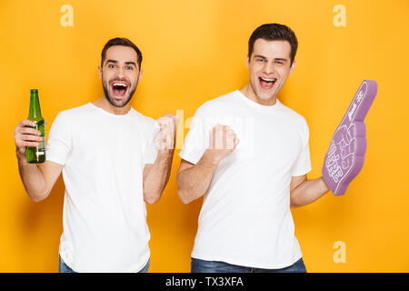 Zwei fröhliche aufgeregt Männer Freunde tragen Leer t-shirts über Gelb Hintergrund isoliert stehen, jubeln mit Schaum Handschuh und Bier Stockfoto