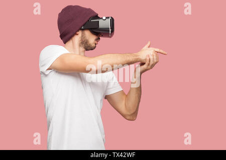 Zuversichtlich bärtigen Jungen hipster Mann im weißen Hemd und casual Hut stehen, tragen vr spielen video game, zeigen, Gewehr singen mit Finger wie Schießen. Stockfoto