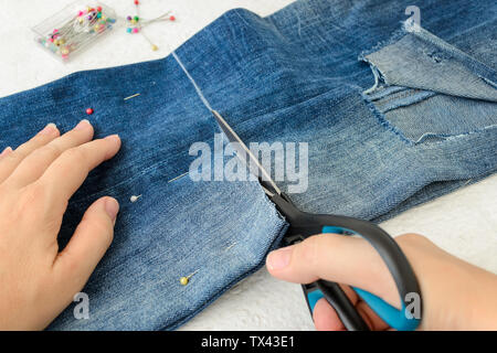 Frau Hand holdig eine Schere und schneiden Sie ein in der Hälfte Blue Jeans mit einem großen Loch auf hosebein gefaltet. Die denim Shorts mit Schere, Stifte und Ta Stockfoto