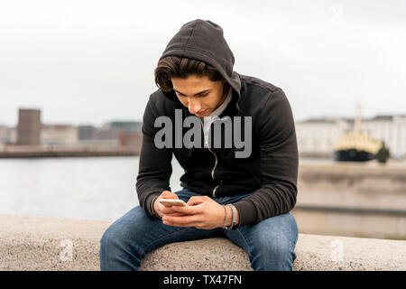 Dänemark, Kopenhagen, junge Mann sitzt auf Wand an der Waterfront können Sie über Handy Stockfoto