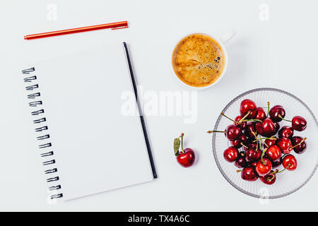 Ansicht von oben Schreibtisch mit weißen leeren Notebook, Tasse Kaffee und die Platte von reifen Kirschen als Snack. Stockfoto