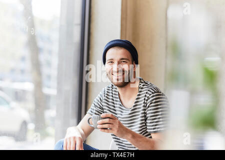 Portrait von lächelnden jungen Mann mit Tasse Kaffee am Fenster
