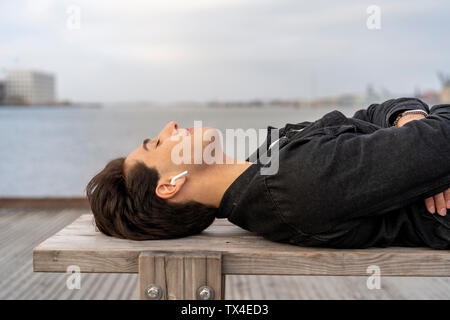 Dänemark, Kopenhagen, junger Mann mit Ohrhörern auf einer Bank an der Uferpromenade liegen Stockfoto