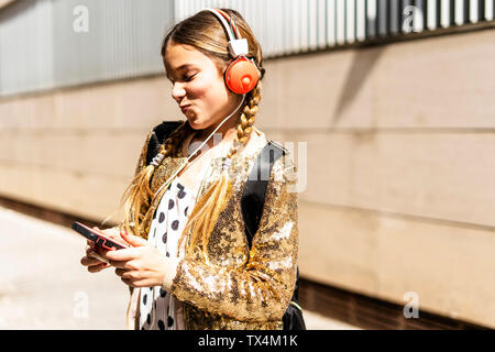 Lächelnde Mädchen mit goldenen Pailletten Jackett und Kopfhörer auf der Suche auf dem Mobiltelefon Stockfoto