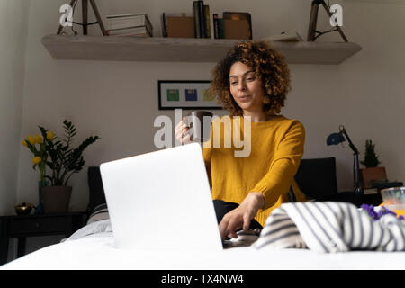 Frau sitzt auf dem Bett, mit Laptop, trinken Kaffee Stockfoto