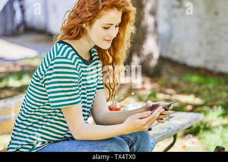 Lächelnd rothaarige junge Frau sitzt auf der Bank im Garten in der Zelle Telefon Stockfoto