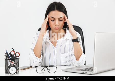 Foto von erschöpft brunette Geschäftsfrau 30 s Tempel reiben beim Sitzen am Tisch mit Laptop im Büro auf weißem Hintergrund Stockfoto