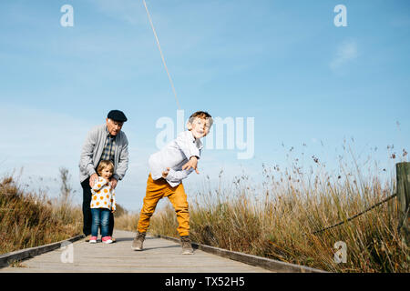 Junge auf Boardwalk werfen Stick, während sein Großvater und seine kleine Schwester ihn aus dem Hintergrund Stockfoto