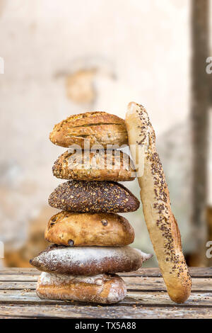 Stapel von mehreren kleinen multi Korn unterschiedlich geformte Brot und Baguette, bestreut mit ganzen Sonnenblumenkerne, Leinsamen und Sesam und Weizen und Gerste spikes, über alte Risse im Lack Wand Stockfoto