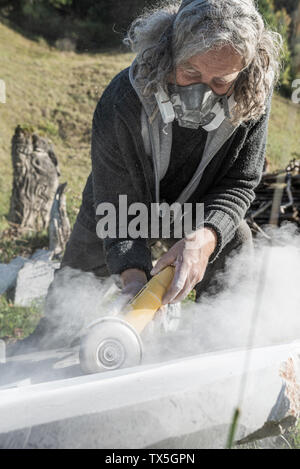 Senior Artist Tragen schützender Dämmerung Maske, während er Polieren ist Stein Skulptur mit Schleifmaschine. Stockfoto