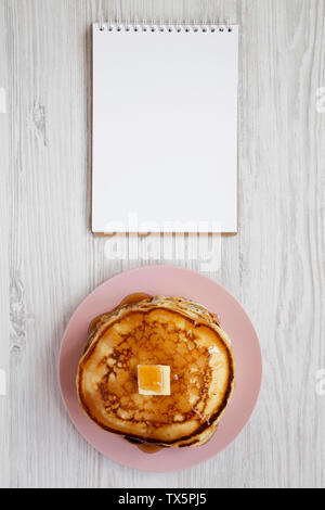 Hausgemachte Pancakes mit Butter und Ahornsirup auf einem rosa Platte, leere Notepad über weiß Holz- Hintergrund, Ansicht von oben. Overhead, flach, von oben. Stockfoto