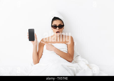 Bild eines schönen überrascht emotionale Frau mit Handtuch auf dem Kopf liegt im Bett unter der Decke über weiße Wand Hintergrund tragen Sonnenbrillen sho Stockfoto