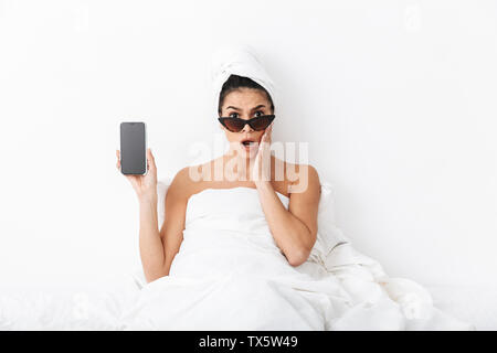 Bild eines schönen überrascht emotionale Frau mit Handtuch auf dem Kopf liegt im Bett unter der Decke über weiße Wand Hintergrund tragen Sonnenbrillen sho Stockfoto