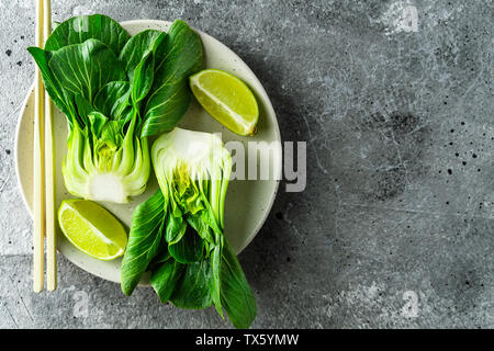 Bok choy Hälften, Kalk Keile, Essstäbchen auf weiße Platte auf grauem Hintergrund. Ansicht von oben mit der Kopie Raum Stockfoto