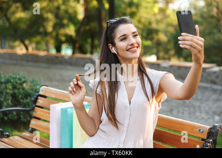 Attraktive junge Frau eine selfie sitzen auf einer Bank im Freien nach dem Einkaufen, Musik hören mit Handy Stockfoto