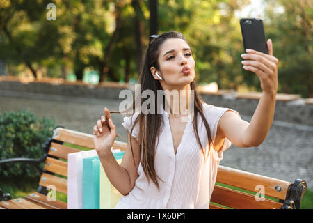 Attraktive junge Frau eine selfie sitzen auf einer Bank im Freien nach dem Einkaufen, Musik hören mit Handy Stockfoto